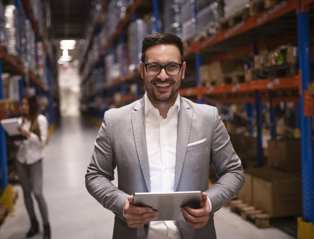 Cheerful and successful middle aged caucasian manager businessman holding tablet computer in large warehouse organizing distribution.